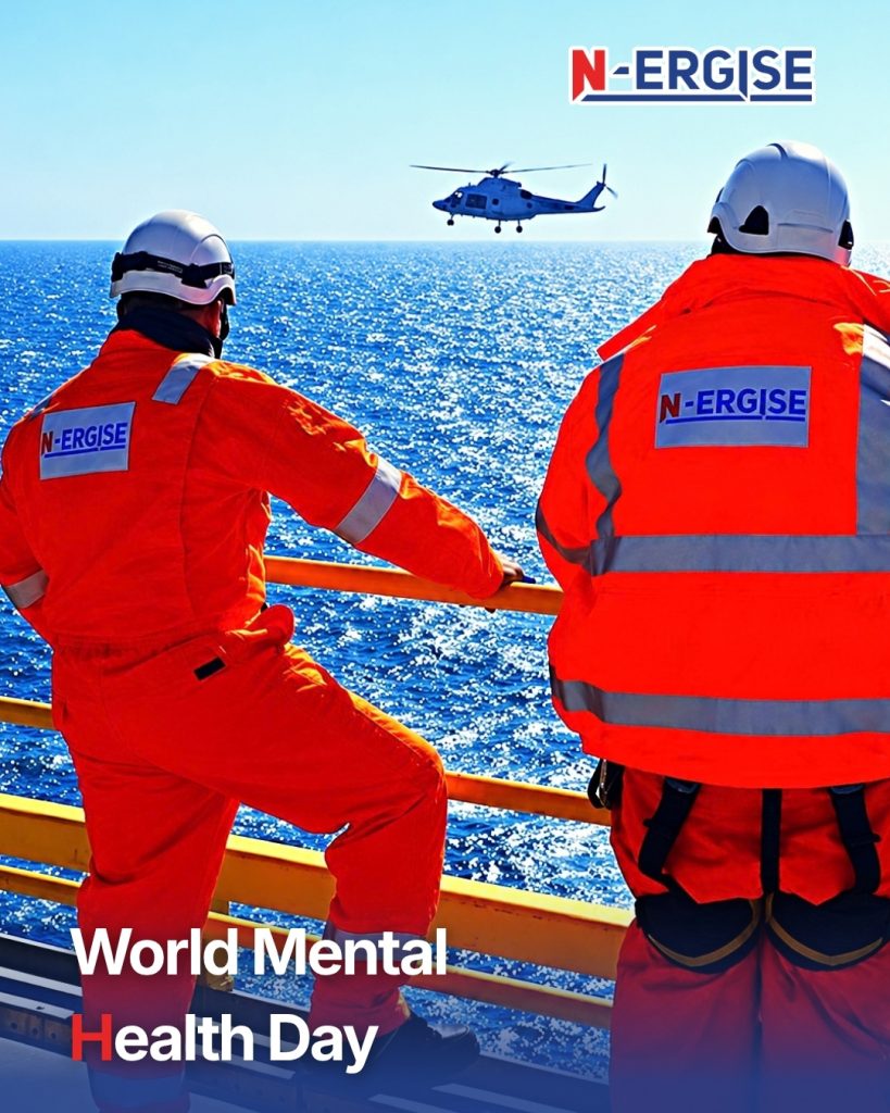 Two N-ERGISE Offshore workers in orange coveralls and white hard hats at a yellow platform rail, watching a helicopter fly over the sea, with the headline World Mental Health Day