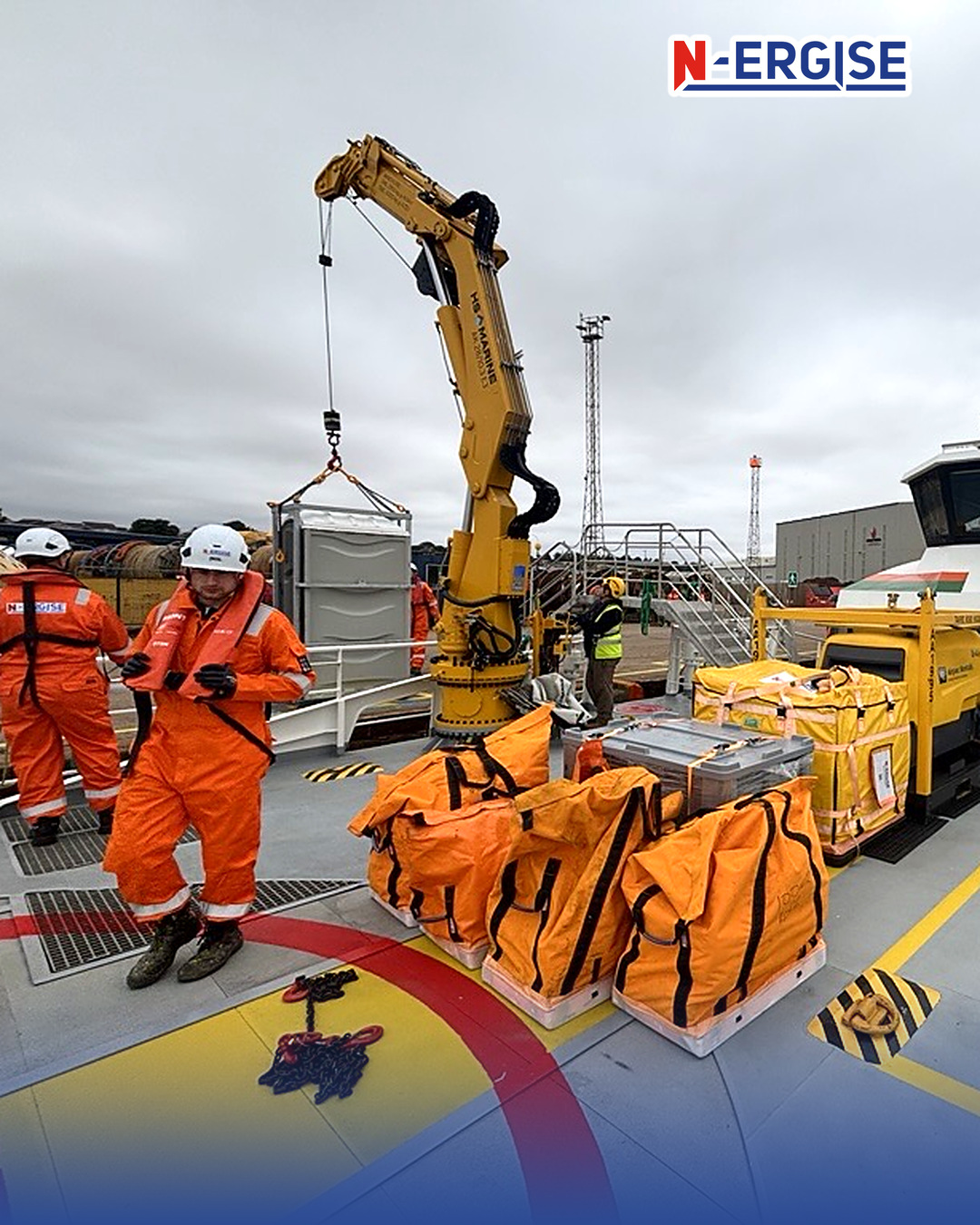 N-ERGISE technicians in orange coveralls and white helmets on a vessel deck, with orange kit bags and equipment boxes being loaded by a yellow deck crane