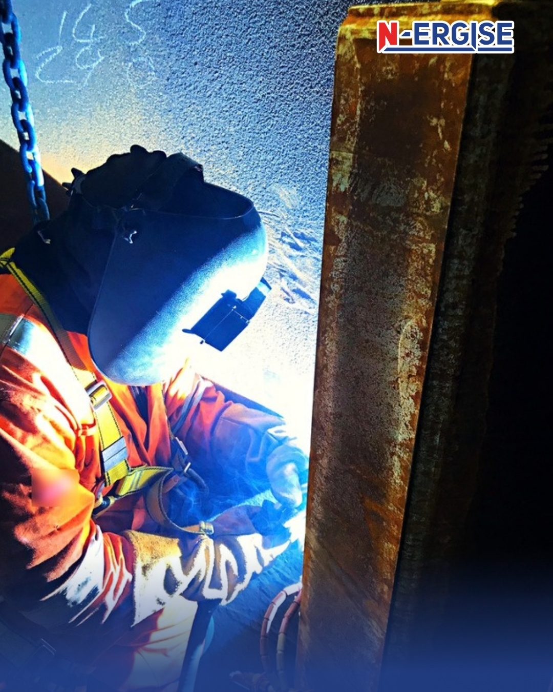 N-ERGISE welder in orange coveralls, safety harness and welding helmet carrying out a weld on a steel shear key inside a structure, with the arc lighting the steel